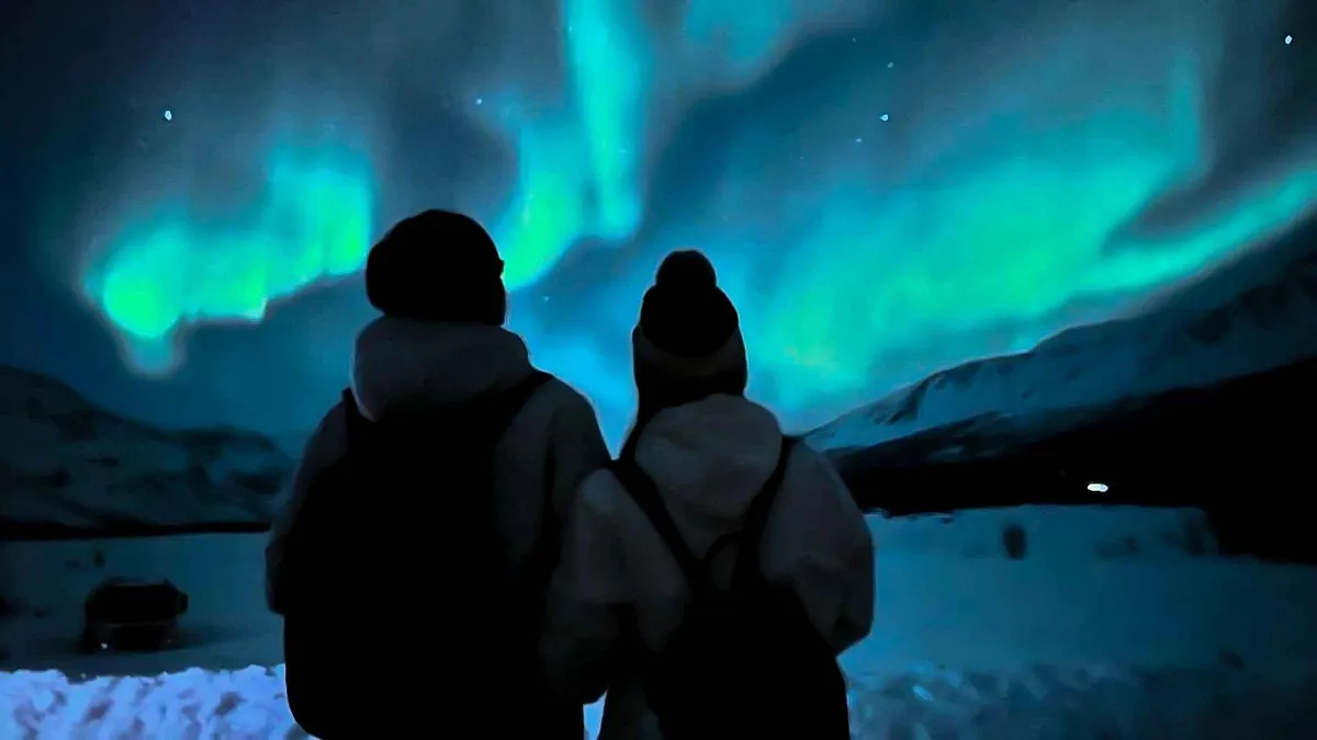 Travellers photographing a green aurora band from a snowy roadside stop on a budget coach Tromso aurora borealis tour, northern Norway